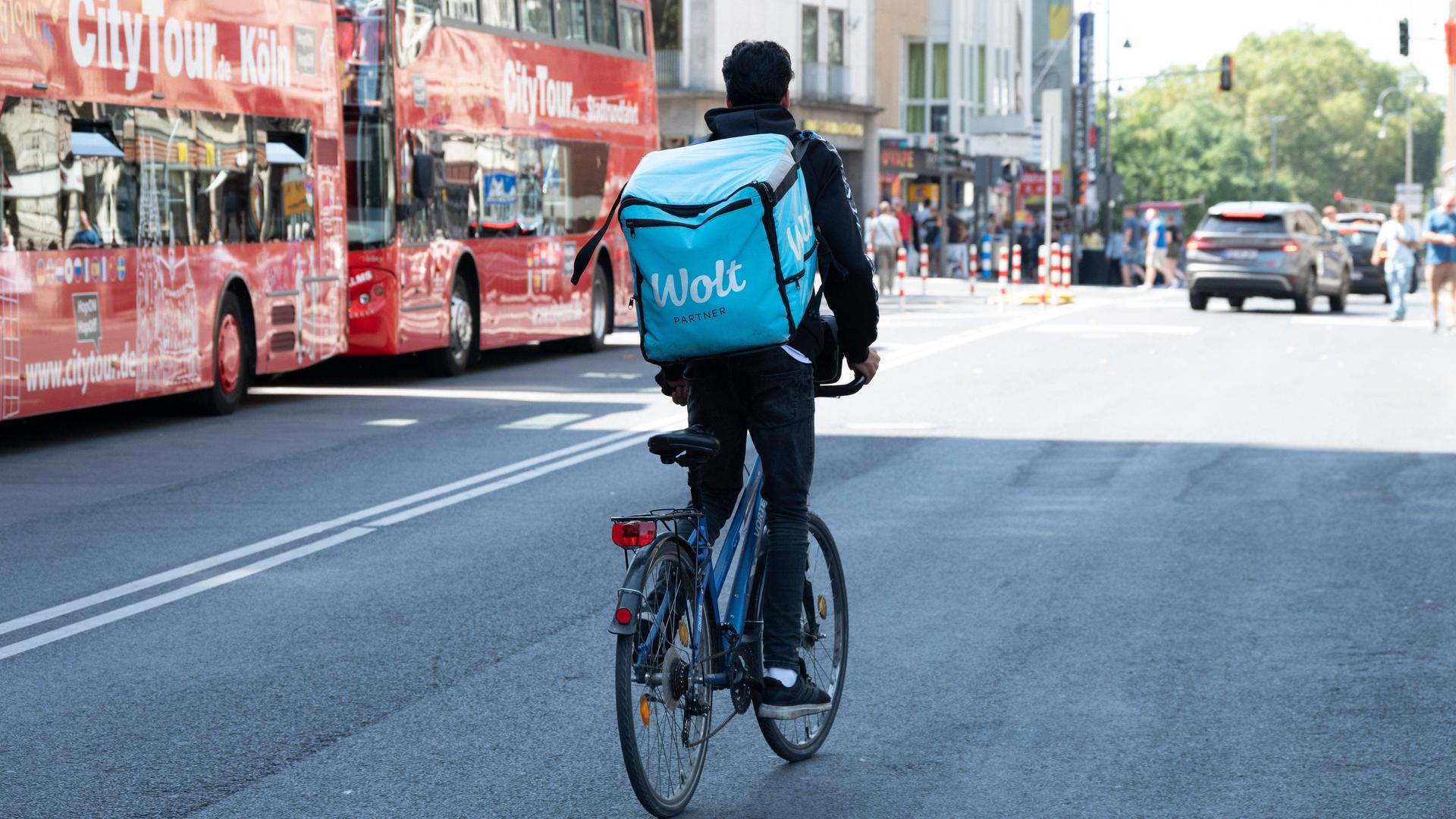 Ein Radfahrer mit einem großen Rucksack auf dem Rücken mit der Aufschrift WOLT fährt auf einer Straße in Köln neben zwei Bussen mit der Aufschrift City Tour 