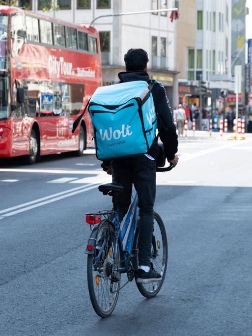 Ein Radfahrer mit einem großen Rucksack auf dem Rücken mit der Aufschrift WOLT fährt auf einer Straße in Köln neben zwei Bussen mit der Aufschrift City Tour 
