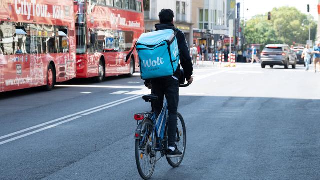 Ein Radfahrer mit einem großen Rucksack auf dem Rücken mit der Aufschrift WOLT fährt auf einer Straße in Köln neben zwei Bussen mit der Aufschrift City Tour 