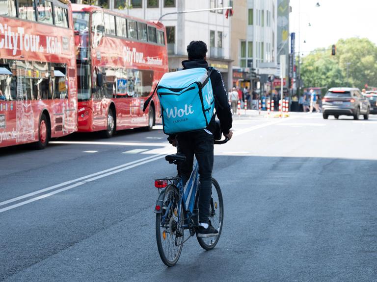 Ein Radfahrer mit einem großen Rucksack auf dem Rücken mit der Aufschrift WOLT fährt auf einer Straße in Köln neben zwei Bussen mit der Aufschrift City Tour 