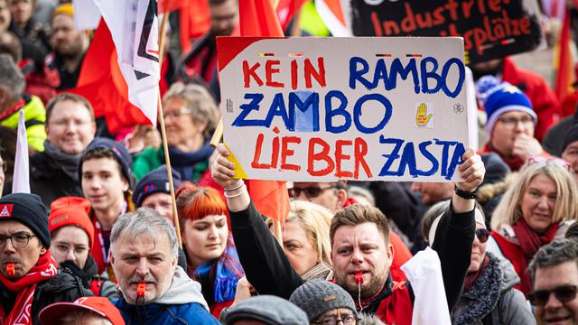 Demonstranten nehmen am bundesweiten Aktionstag der IG Metall teil. Einer hält ein Schild hoch mit der Aufschrift: "Kein Rambo Zambo, lieber Zasta".