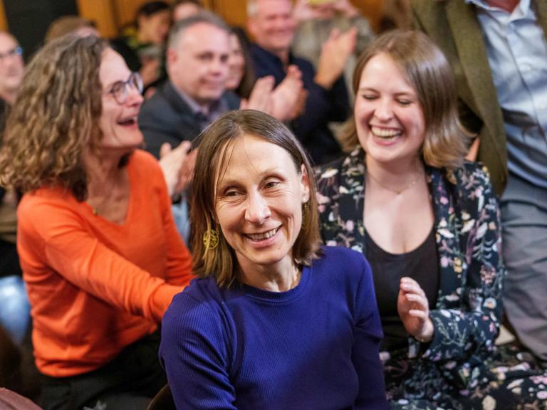 Martina Hefter erhebt sich lachend aus dem Publikum bei der Preisverleihung, die Menschen um sie herum applaudieren