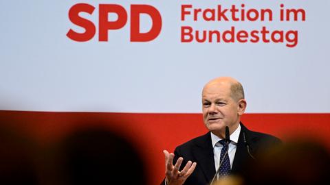 Bundeskanzler Scholz spricht bei einer Veranstaltung und hebt dabei seine rechte Hand, die Finger zeigen nach oben.