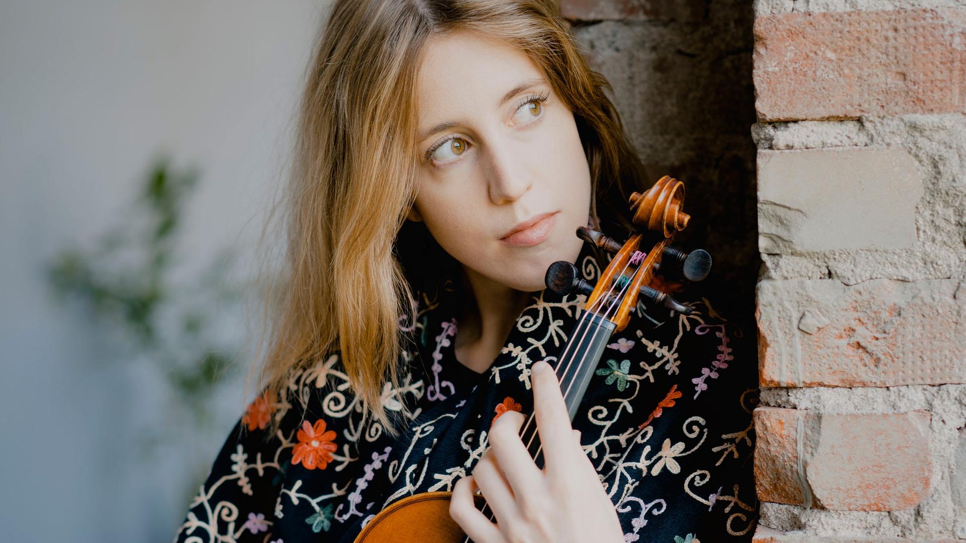 Vilde Frang steht mit einer Bluse voller Blumen nachdenklich an eine Ziegelmauer gelehnt, während sie ihre Geige umschließt.