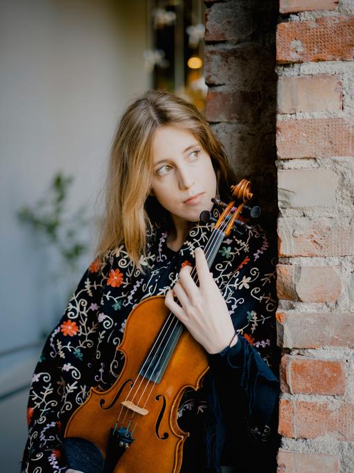 Vilde Frang steht mit einer Bluse voller Blumen nachdenklich an eine Ziegelmauer gelehnt, während sie ihre Geige umschließt.