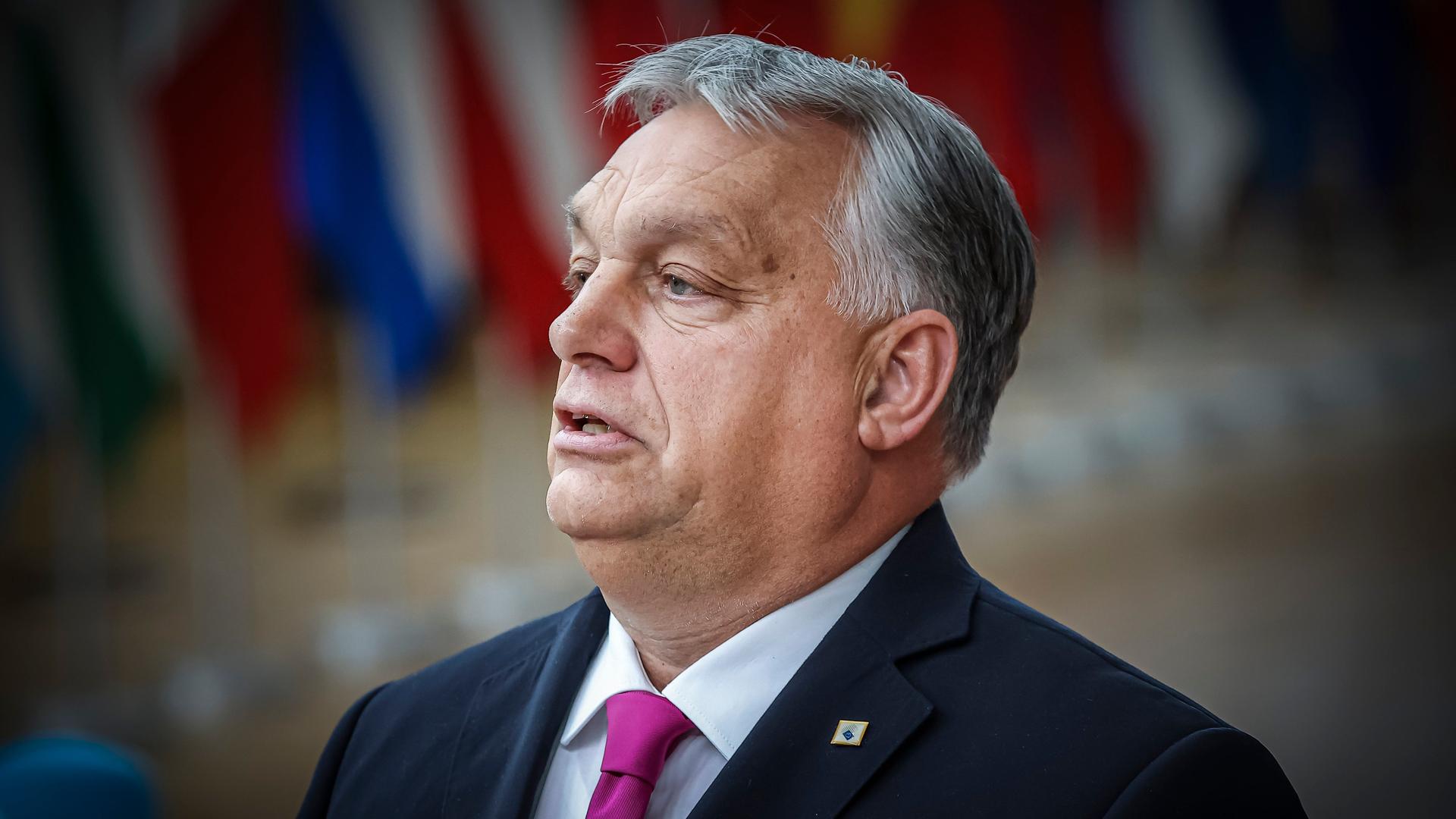 Ungarns Premierminister Viktor Orbán steht in Brüssel, im Hintergrund sind unscharf Flaggen der EU-Staaten zu sehen. 