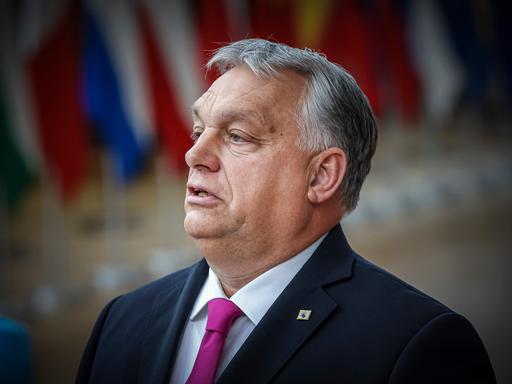 Ungarns Premierminister Viktor Orbán steht in Brüssel, im Hintergrund sind unscharf Flaggen der EU-Staaten zu sehen. 