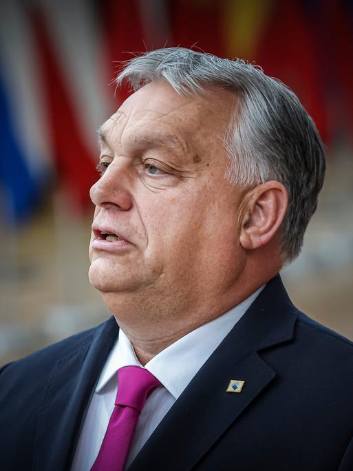 Ungarns Premierminister Viktor Orbán steht in Brüssel, im Hintergrund sind unscharf Flaggen der EU-Staaten zu sehen. 