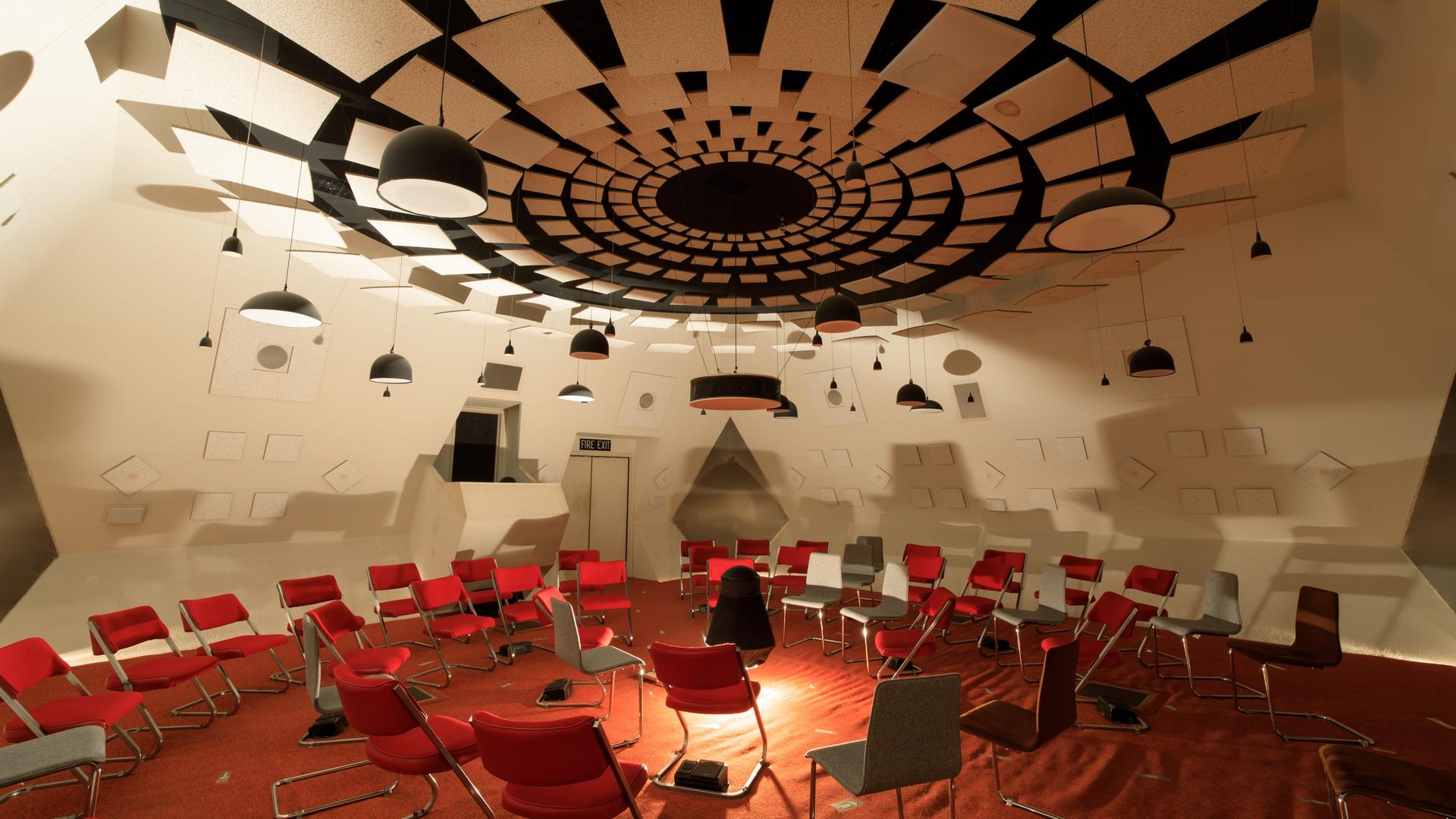 Ein futuristisch anmutender Raum in weiß mit roten Stühlen und den unterschiedlichsten hängenden, stehenden und verbauten Lautsprechern Ein futuristisch anmutender Raum in weiß mit roten Stühlen und den unterschiedlichsten hängenden, stehenden und verbauten Lautsprechern