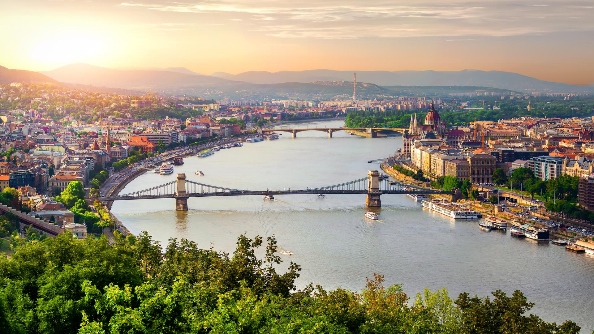 Blick über das Zentrum von Budapest mit zwei Brücken über die Donau. Am Horizont ist ein Sonnenaufgang zu sehen.