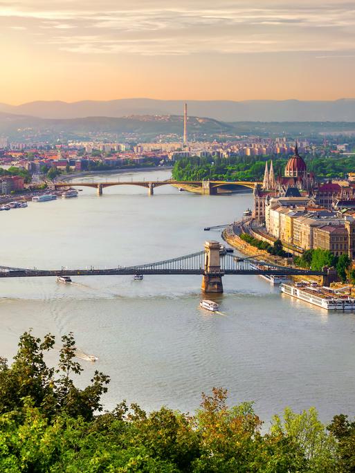 Blick über das Zentrum von Budapest mit zwei Brücken über die Donau. Am Horizont ist ein Sonnenaufgang zu sehen.