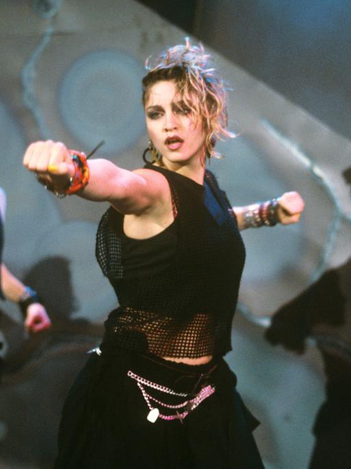 Madonna auf der Bühne, aufgenommen im März 1984 in München. Im Hintergrund eine Tänzerin und ein Tänzer. Alle tragen schwarz. Die rechte Faust ist nach vorne gestreckt.