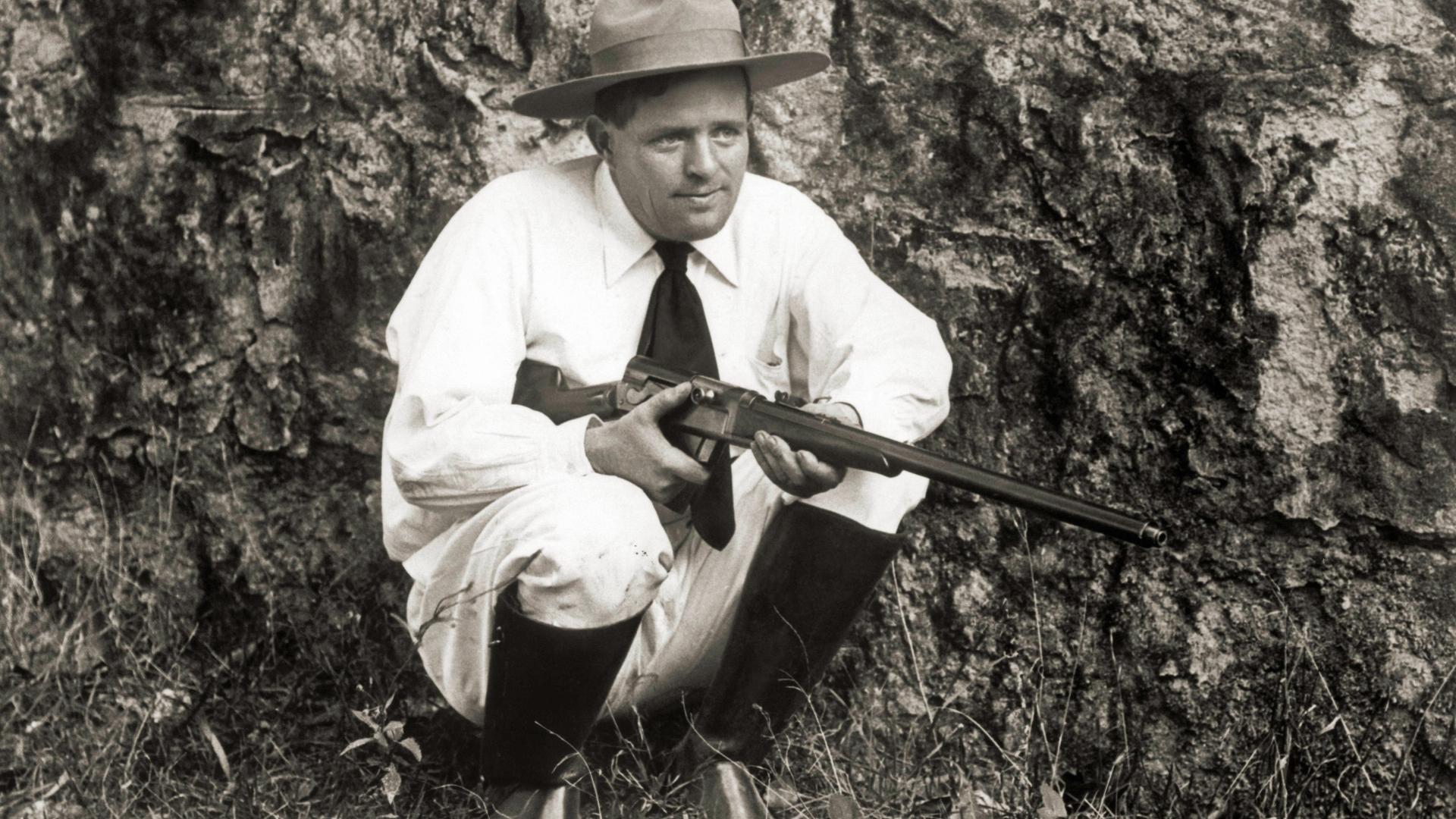 Der Schriftsteller Jack London kniet mit einer Flinte im Gras