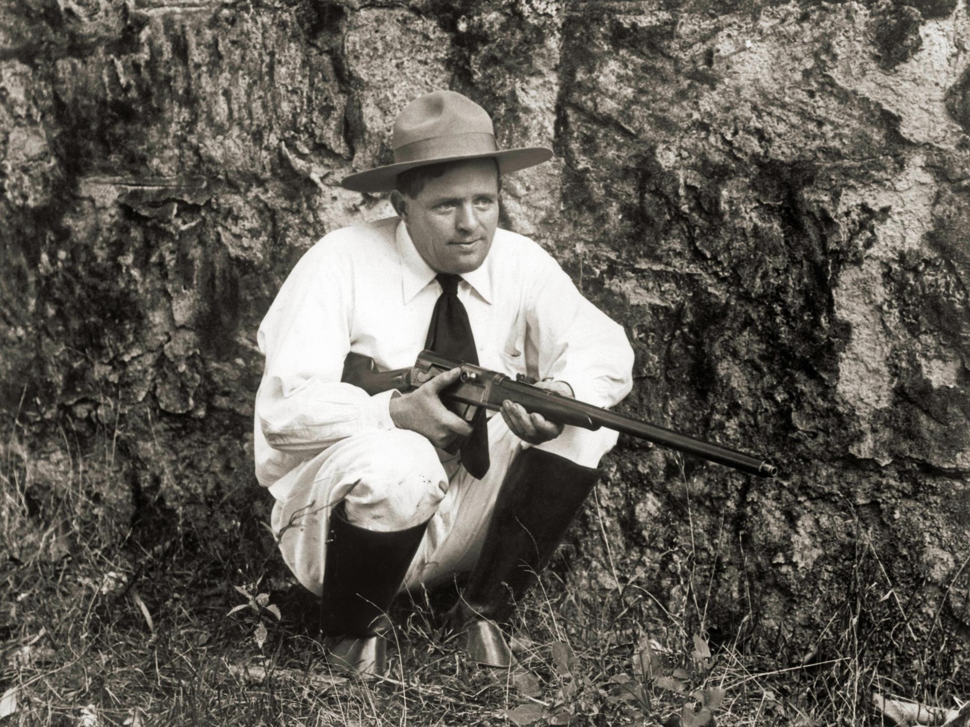 Der Schriftsteller Jack London kniet mit einer Flinte im Gras