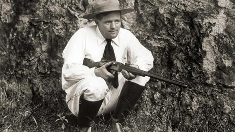 Der Schriftsteller Jack London kniet mit einer Flinte im Gras
