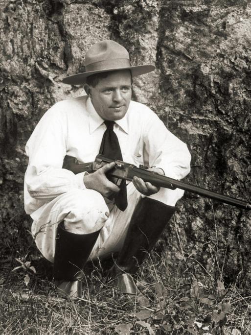 Der Schriftsteller Jack London kniet mit einer Flinte im Gras