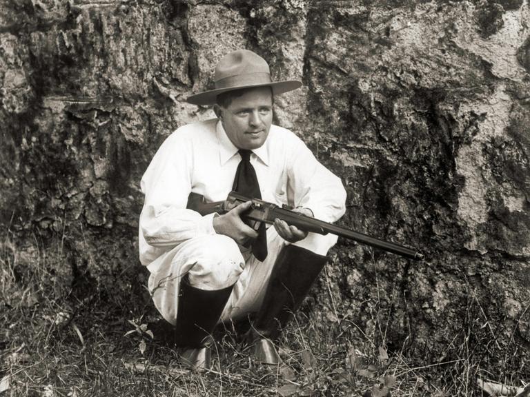 Der Schriftsteller Jack London kniet mit einer Flinte im Gras