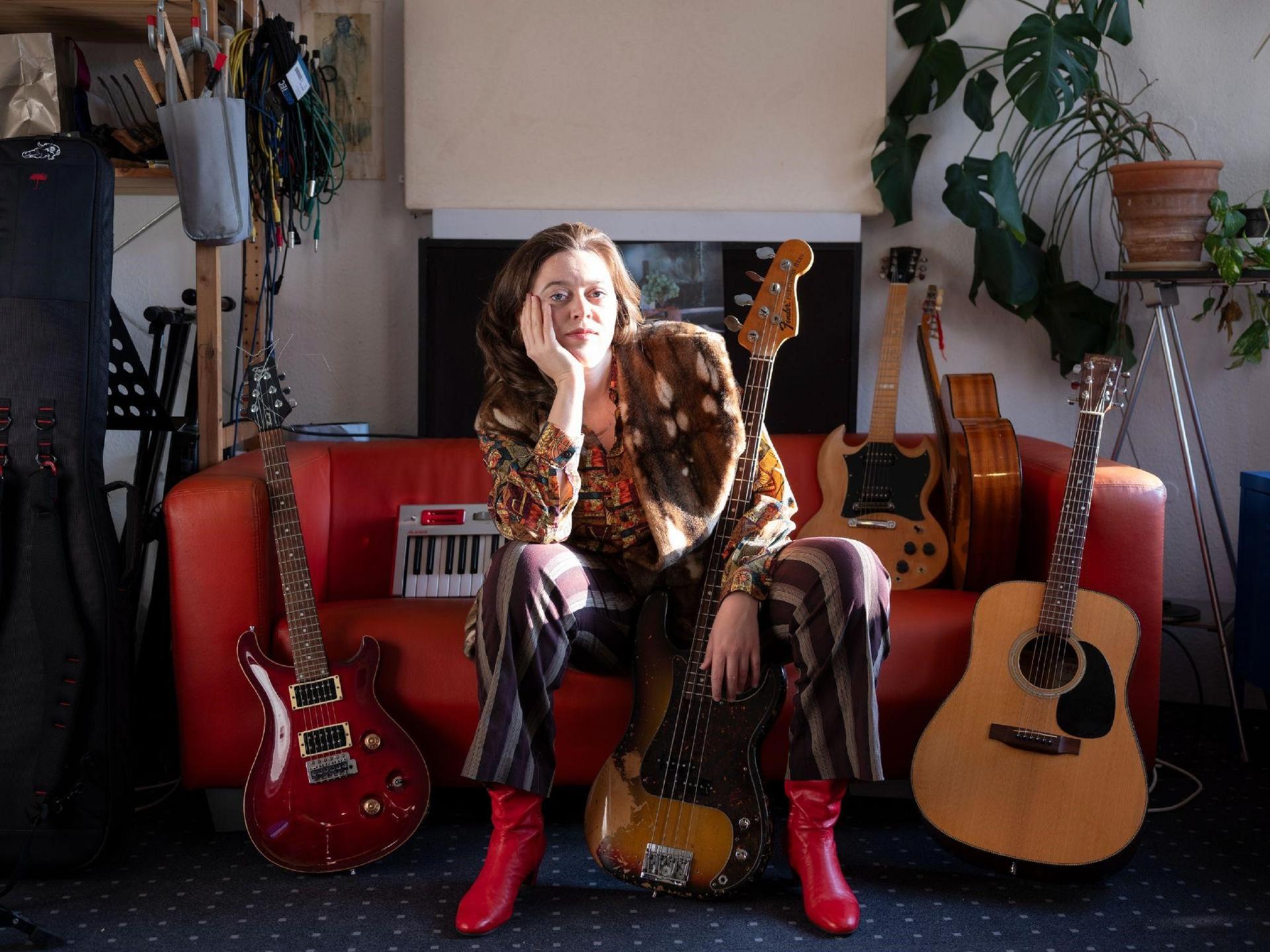 Eine Frau sitzt mit roten Stiefeln auf einem roten Ledersofa, auf dem viele Gitarren aufgebaut sind.