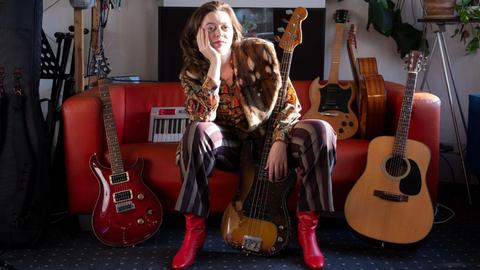 Eine Frau sitzt mit roten Stiefeln auf einem roten Ledersofa, auf dem viele Gitarren aufgebaut sind.