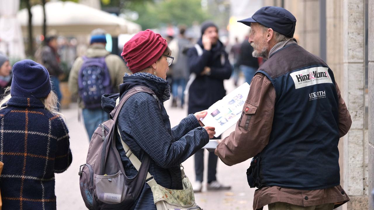 Ein Mann mit "Hinz&Kunzt"-Weste reicht einer Frau in einer Fußgängerzone eine Papierausgabe