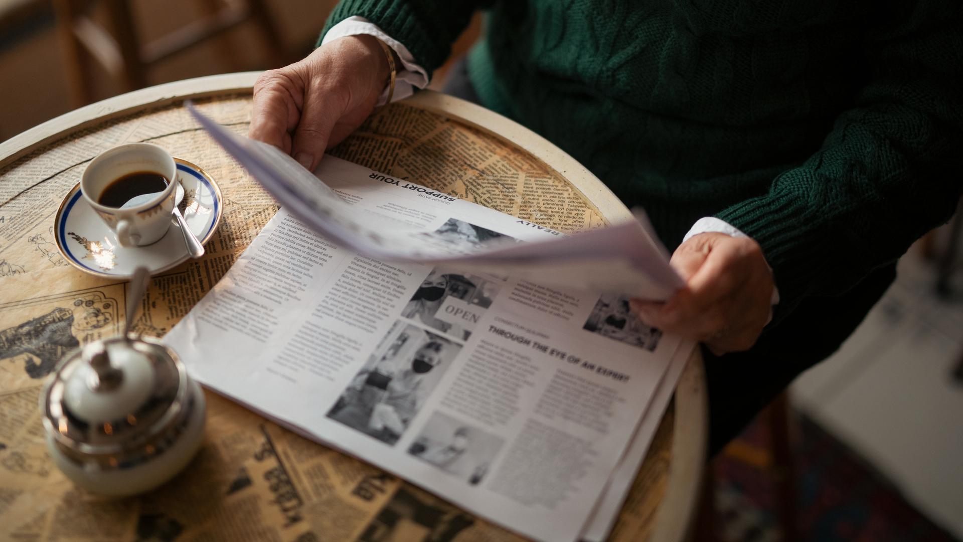Ein älterer Mensch im Ausschnitt sitzt mit einer Zeitung an einem Tisch in einem Café und trinkt Kaffee