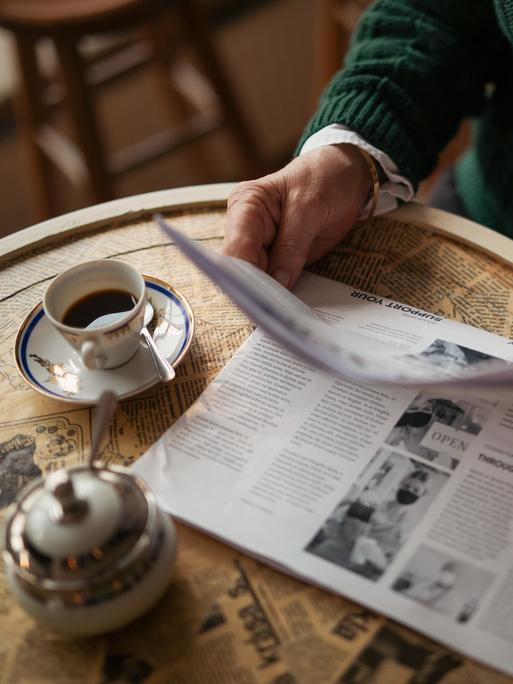 Eine älterer Mensch im Ausschnitt sitzt mit einer Zeitung an einem Tisch in einem Café und trinkt Kaffee