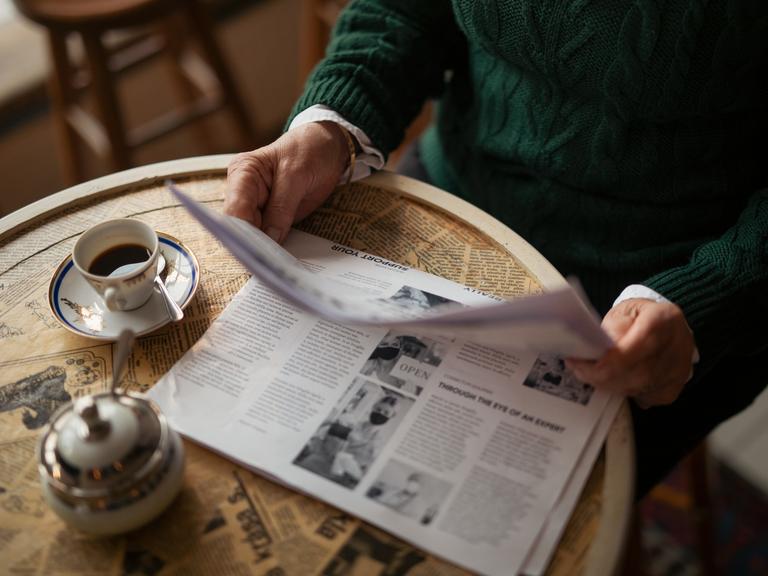 Eine älterer Mensch im Ausschnitt sitzt mit einer Zeitung an einem Tisch in einem Café und trinkt Kaffee