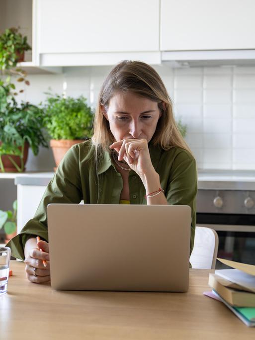 Eine Frau sitzt am Küchentisch an ihrem Laptop; neben ihr ein Handy, ein Wasserglas und ein Stapel Hefte und Bücher. 