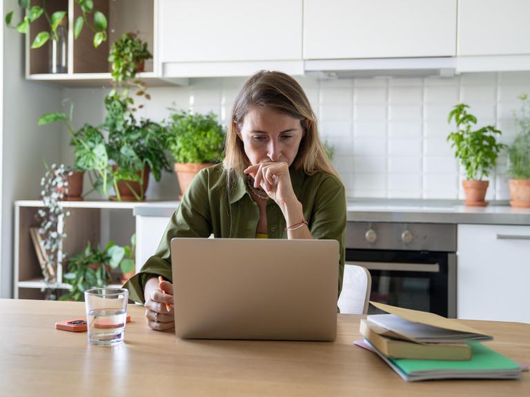 Eine Frau sitzt am Küchentisch an ihrem Laptop; neben ihr ein Handy, ein Wasserglas und ein Stapel Hefte und Bücher. 