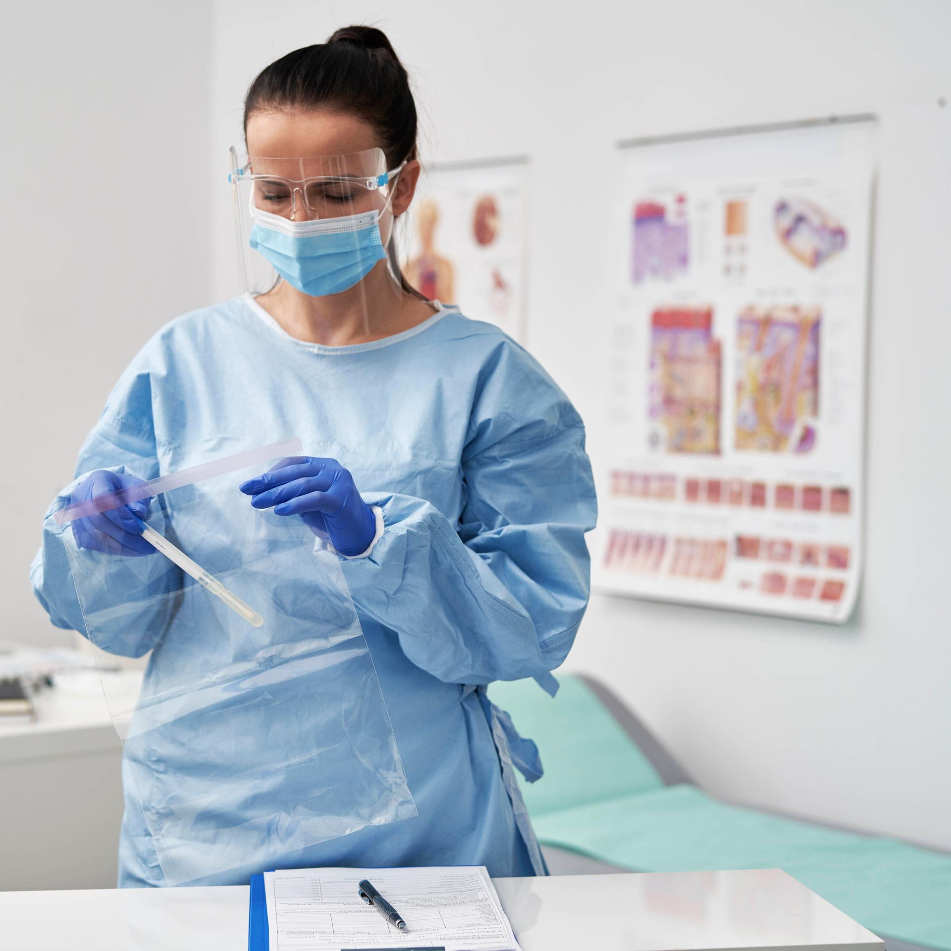 Eine Ärztin im hellblauen Kittel und mit Mundschutz in ihrem Sprechzimmer. Sie packt etwas in eine Plastikhülle.