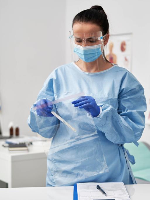 Eine Ärztin im hellblauen Kittel und mit Mundschutz in ihrem Sprechzimmer. Sie packt etwas in eine Plastikhülle.