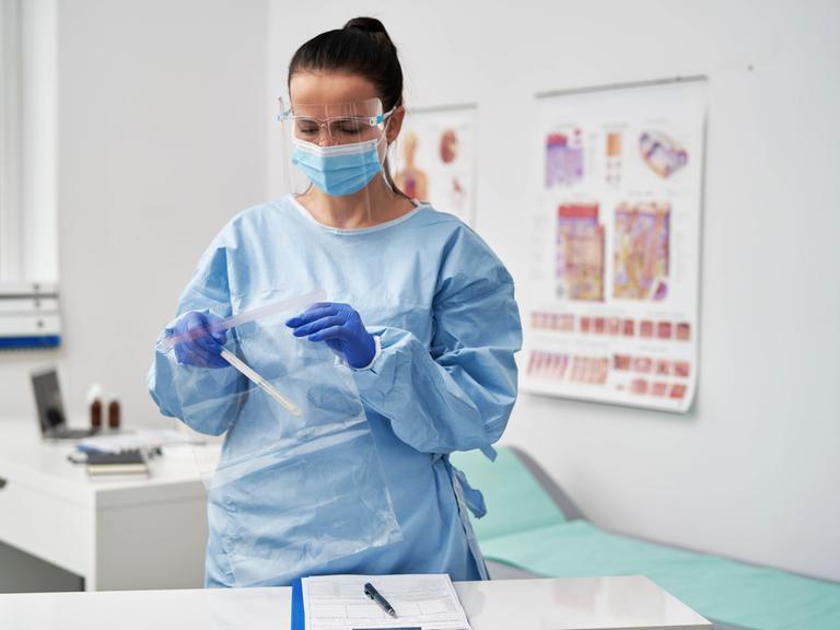 Eine Ärztin im hellblauen Kittel und mit Mundschutz in ihrem Sprechzimmer. Sie packt etwas in eine Plastikhülle.