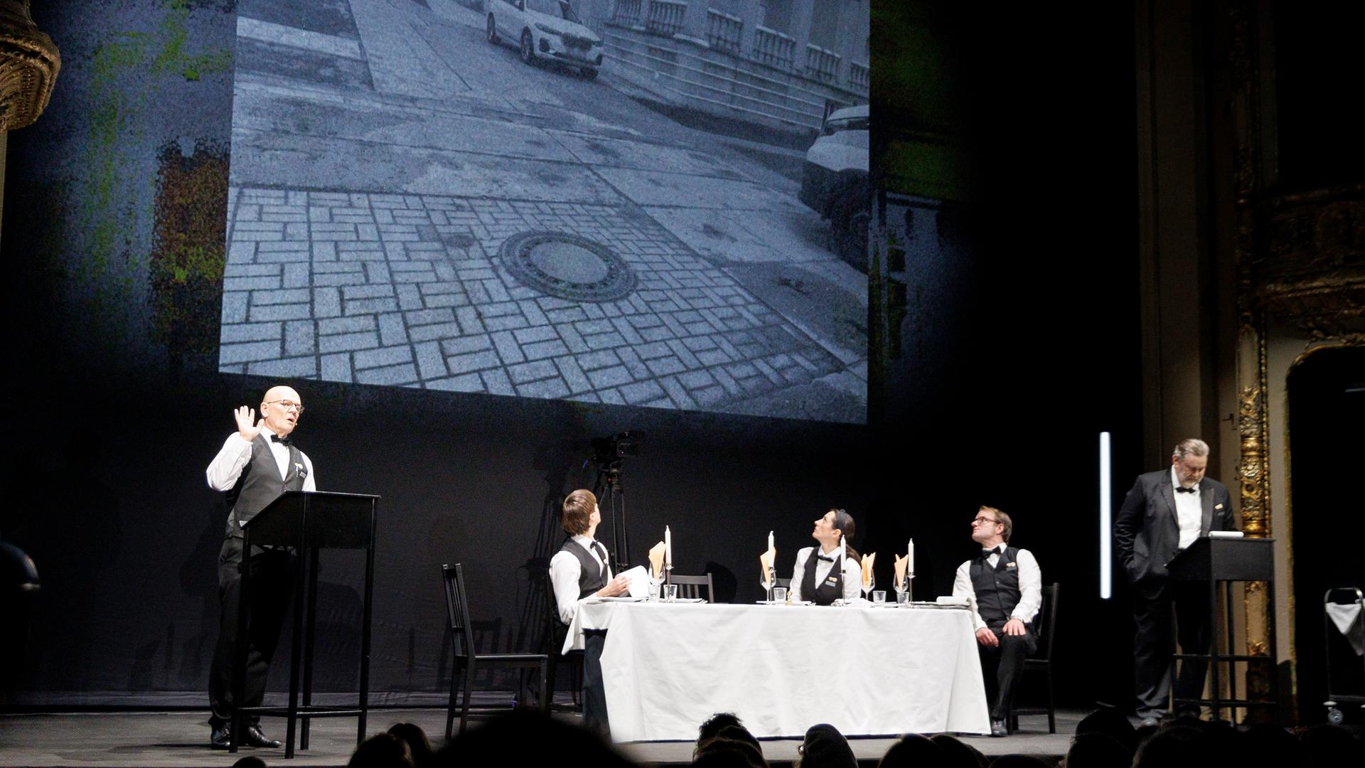 Vier Schauspieler und eine Schauspielerin in Kellner-Kostümen agieren an einer Tafel mit weißem Tischtuch und an zwei Stehpulten.