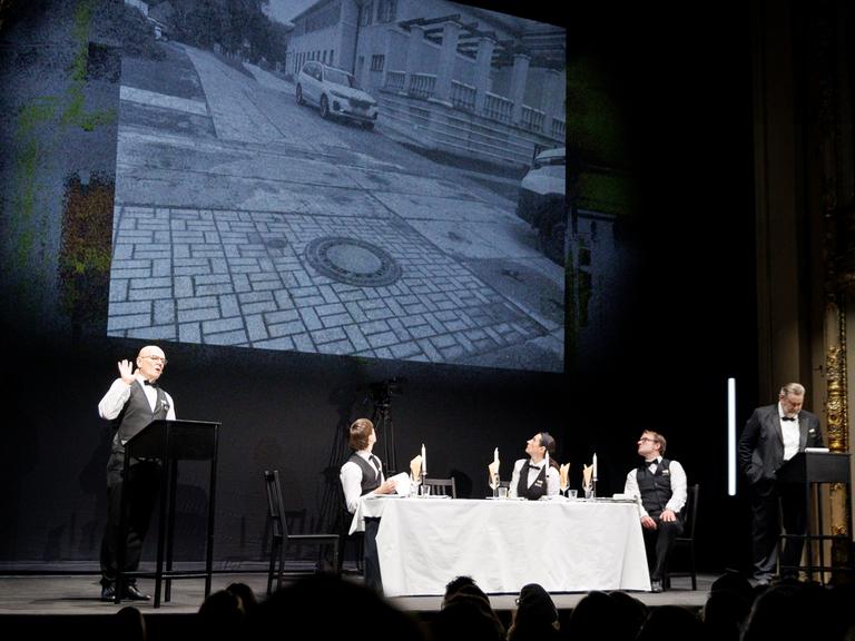 Vier Schauspieler und eine Schauspielerin in Kellner-Kostümen agieren an einer Tafel mit weißem Tischtuch und an zwei Stehpulten.