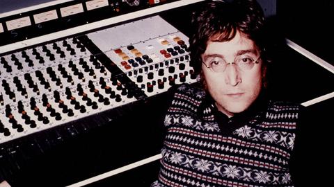 John Lennon sitzt an einer Studiokonsole aus den Siebzigern und blickt ernst in die Kamera