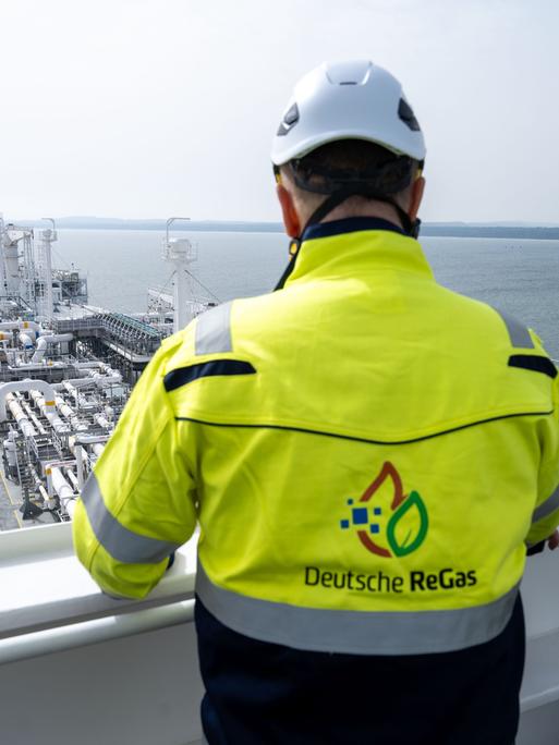 Ein Mann mit Helm und Jacke mit der Aufschrift "Deutsche ReGas" steht auf der Brücke des Speicherschiffs "Energos Power" im Hafen von Mukran. 