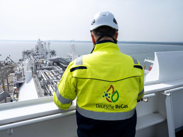 Ein Mann mit Helm und Jacke mit der Aufschrift "Deutsche ReGas" steht auf der Brücke des Speicherschiffs "Energos Power" im Hafen von Mukran. 