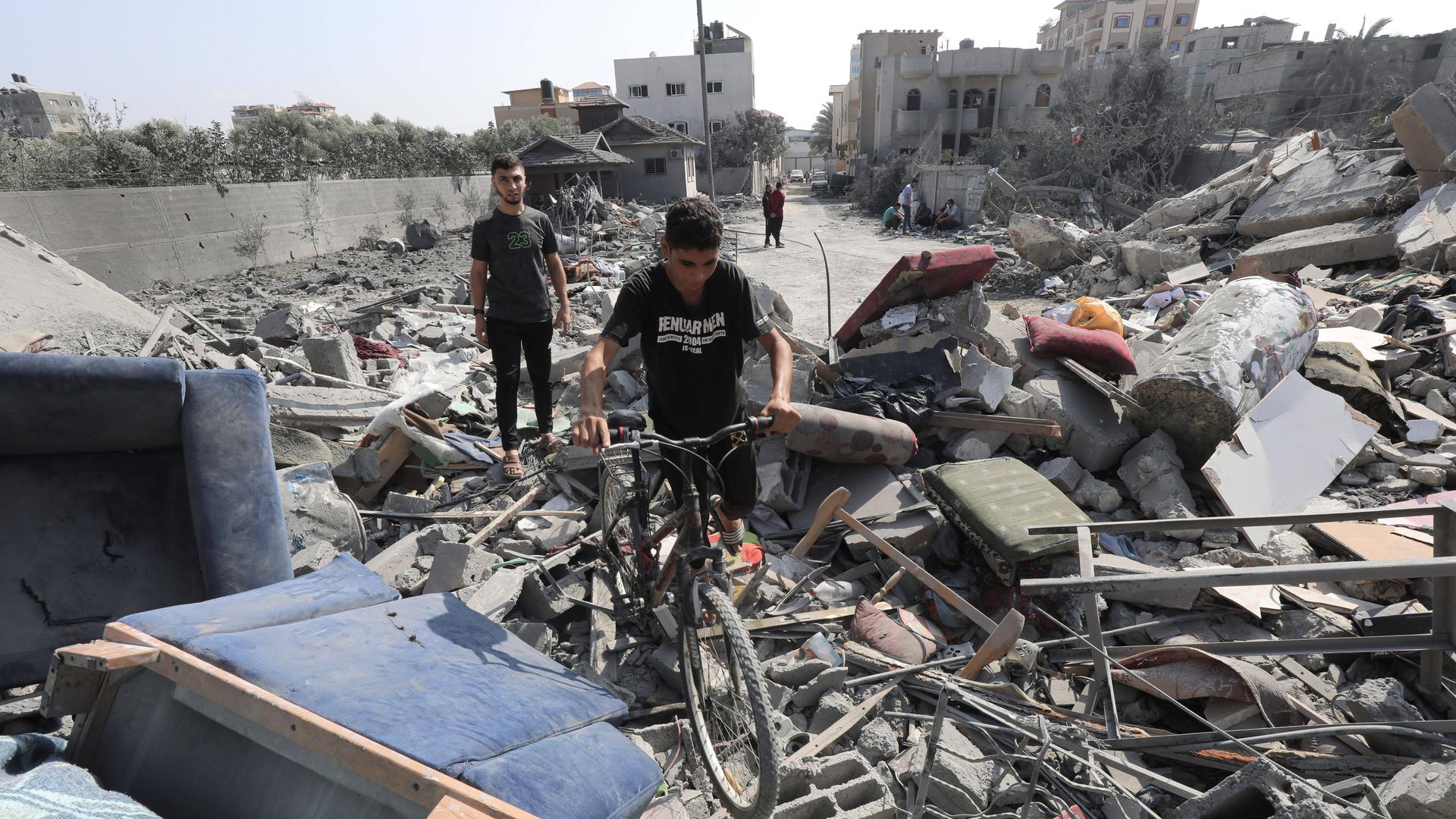 Ein Junge schiebt sein Fahrrad über Trümmer eines Hauses im Gazastreifen, im Hintergrund steht ein Mann.