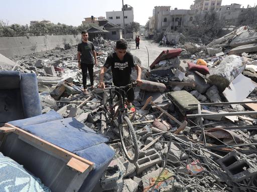Ein Junge schiebt sein Fahrrad über Trümmer eines Hauses im Gazastreifen, im Hintergrund steht ein Mann.