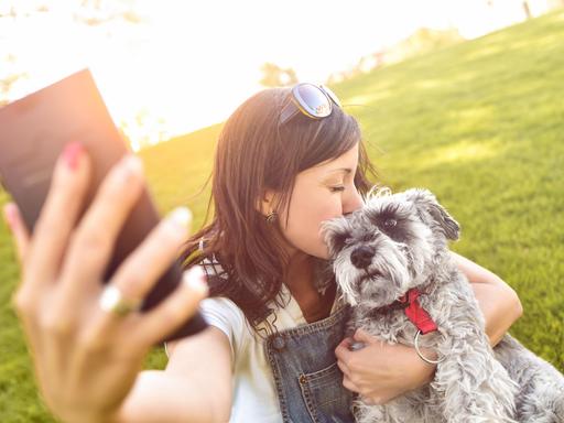Eine junge Frau hält einen Hund im Arm und macht ein Foto mit einem Smartphone.