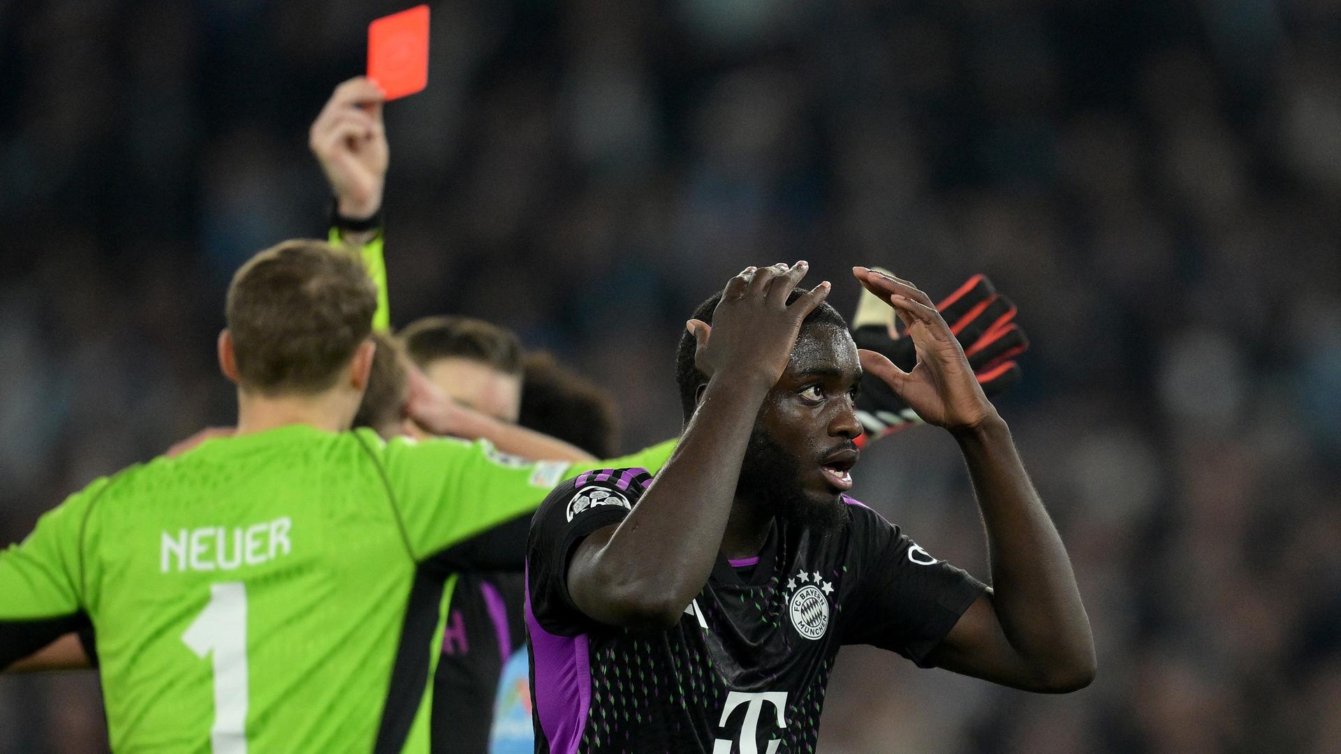 Upamecano im schwarzen Trikot hält bestürzt die Hände an den Kopf. Torhüter Neuer sieht man von hinten protestieren. Dahinter unscharf der erhobene Arm des Schiedsrichters mit der Roten Karte.