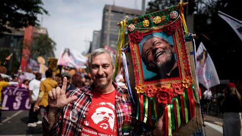 Ein Mann steht in einer Menge auf der Straße und hält ein großes buntumrandetes Foto von Lula da Silva an einem Stock in der linken Hand. Mit der rechten streckt er Zeigefinger und Daumen in die Höhe.