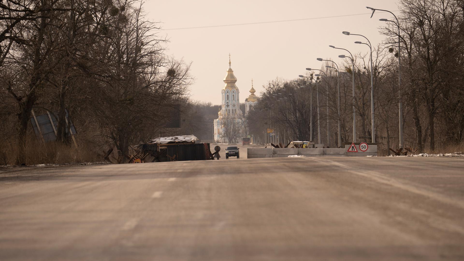 Blick von weitem auf den Checkpoint auf einer breiten, von Bäumen gesäumte Straße, durch den gerade ein Auto fährt. Im Hintergrund eine orthodoxe Kirche.