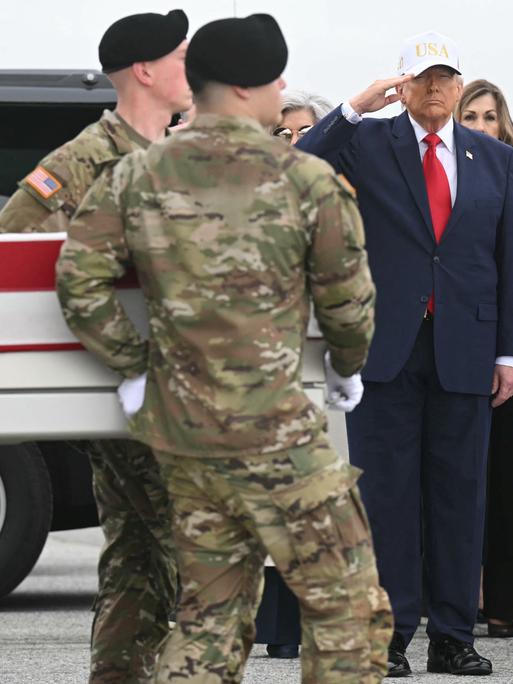 Donald Trump salutiert, während Soldaten einen in die US-Flagge gehüllten Sarg an ihm vorbeitragen.