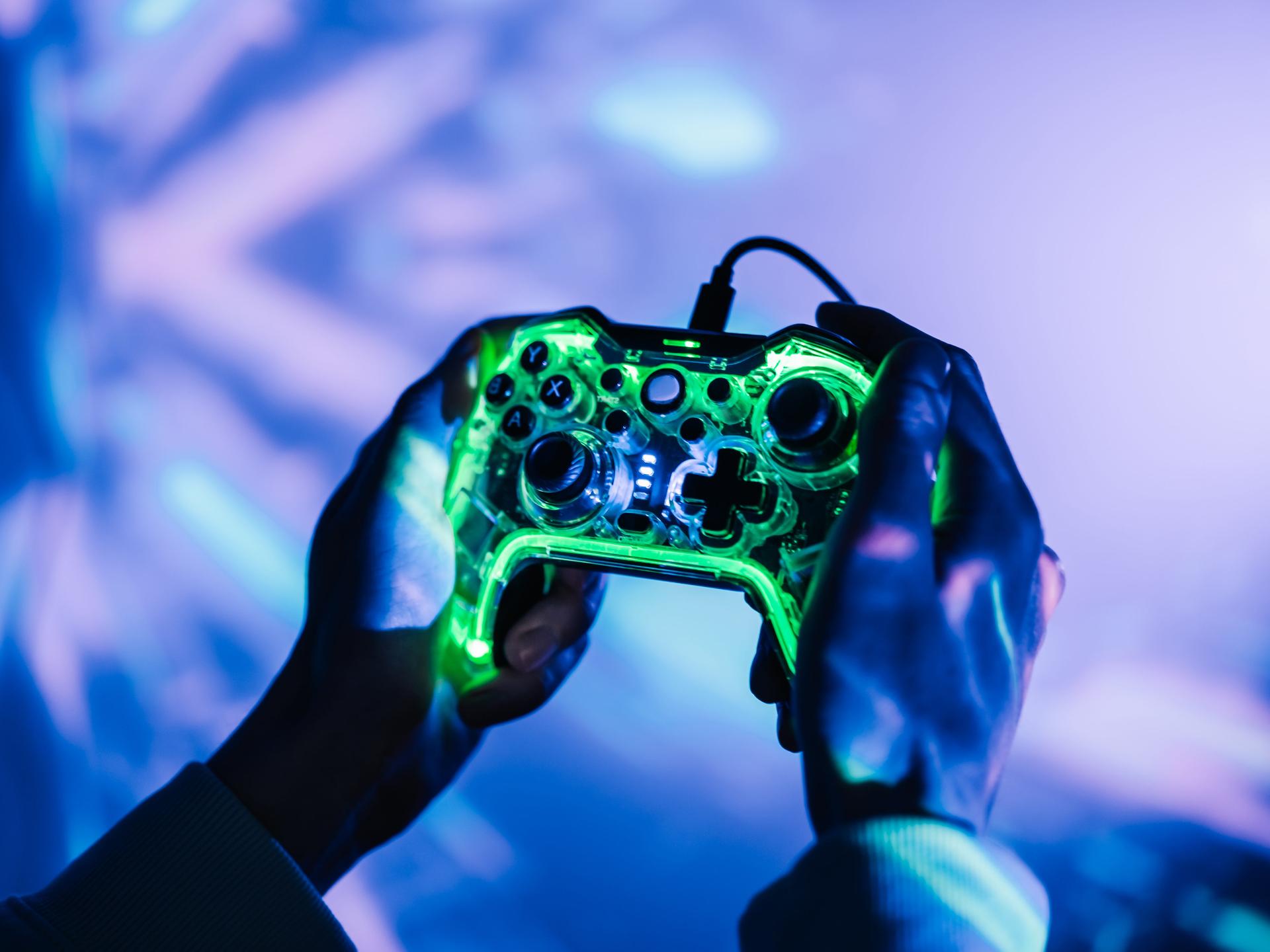 Nahaufnahme zweier Hände die eine Gamingkonsole halten. Die Umgebung ist geprägt von neonfarbenen Lichtern.