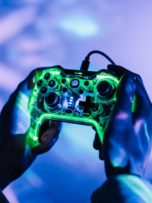 Nahaufnahme zweier Hände die eine Gamingkonsole halten. Die Umgebung ist geprägt von neonfarbenen Lichtern.