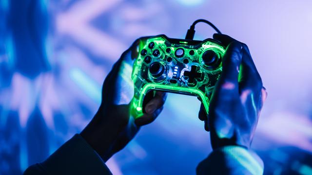 Nahaufnahme zweier Hände die eine Gamingkonsole halten. Die Umgebung ist geprägt von neonfarbenen Lichtern. Nahaufnahme zweier Hände die eine Gamingkonsole halten. Die Umgebung ist geprägt von neonfarbenen Lichtern.
