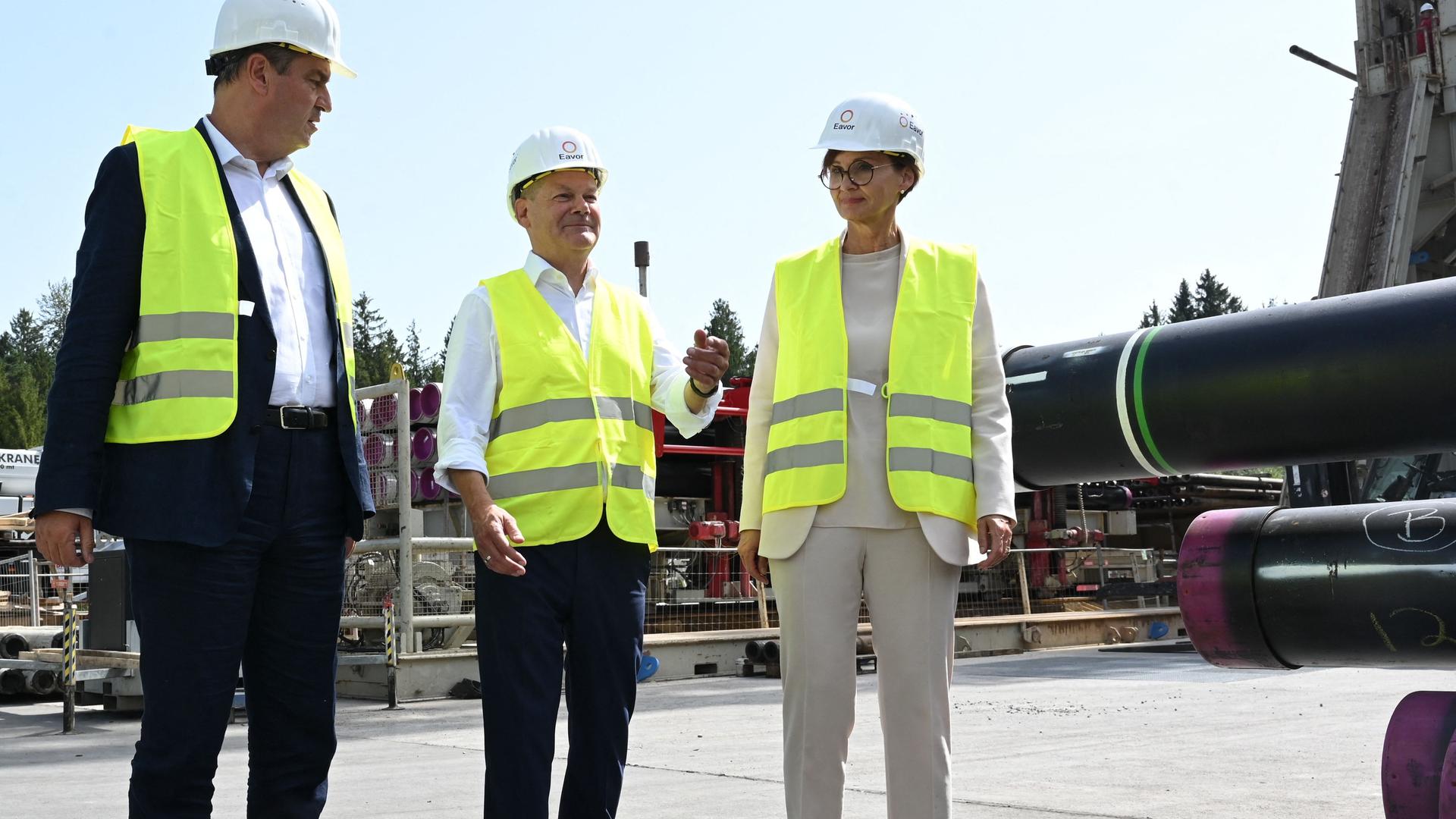 Bundeskanzler Scholz, der bayerische Ministerpräsident Söder und Bundesbildungsministerin Stark-Watzinger zu Besuch in einem Tiefen-Geothermiekraftwerk in Geretsried in Bayern. Sie tragen gelbe Sicherheitswesten und einen Schutzhelm.