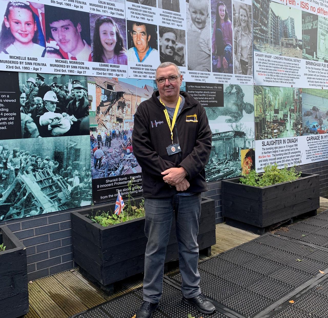 Frank steht vor einer Fotowand mit Opfern der IRA-Gewalt in Nordirland.