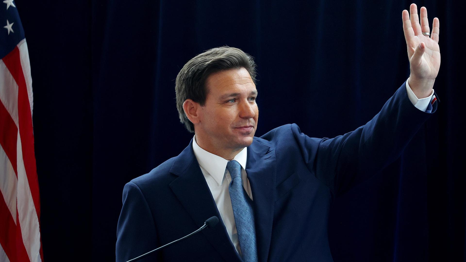 DeSantis mit dunklem Anzug und Krawatte winkt neben einer US-Fahne mit der linken Hand. Der Hintergrund ist dunkel. DeSantis mit dunklem Anzug und Krawatte winkt neben einer US-Fahne mit der linken Hand. Der Hintergrund ist dunkel.